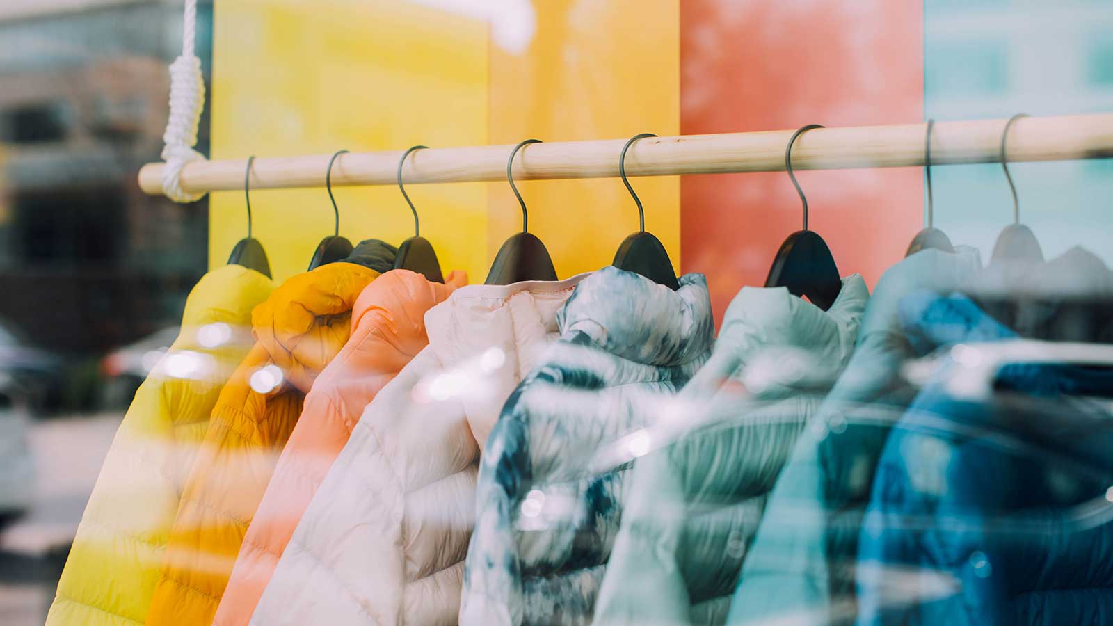 Colorful puffer jackets hang on black hangers from a wooden rack in a store window display at Gardens Centre, with a yellow and orange background and reflections on the glass.