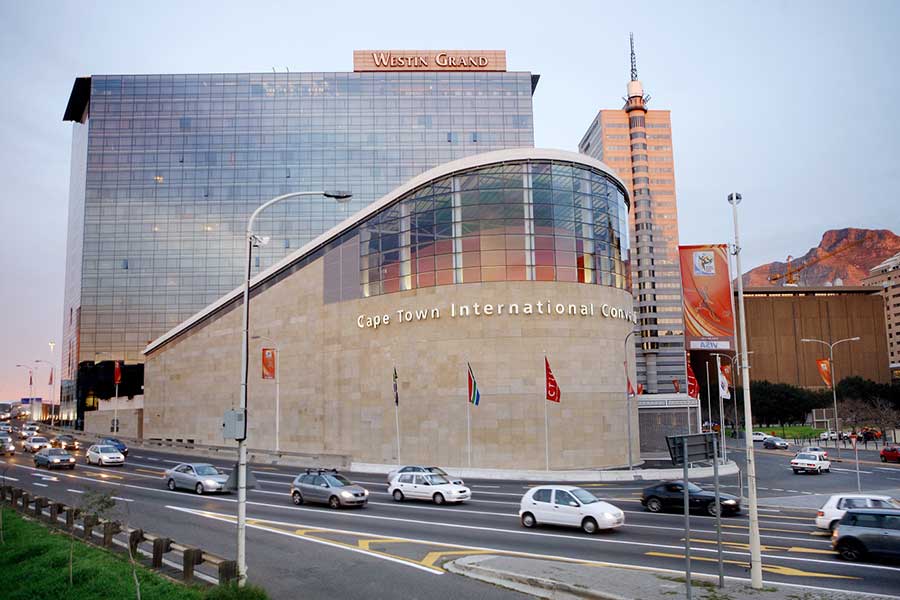 A busy street with cars and buses passes in front of the modern glass and stone exterior of the Cape Town International Convention Centre, with the Westin Grand hotel and city buildings in Cape Town visible in the background.