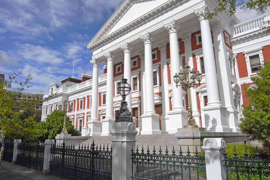 A grand neoclassical building with tall white columns and red brick walls, the South African Parliament stands surrounded by greenery and a black iron fence under a partly cloudy sky.